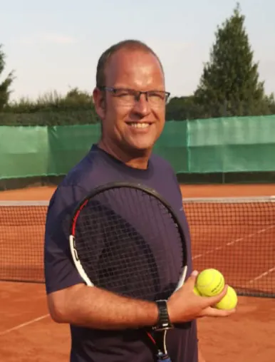 Dino Gulcz steht auf einen Asche-Tennisplatz mit Tennisbällen in der Hand und seinem Tennisschläger unterm Arm.